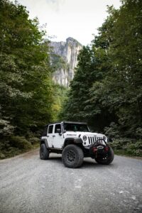 A rugged Jeep Wrangler on a gravel road surrounded by lush forest and towering mountains.
